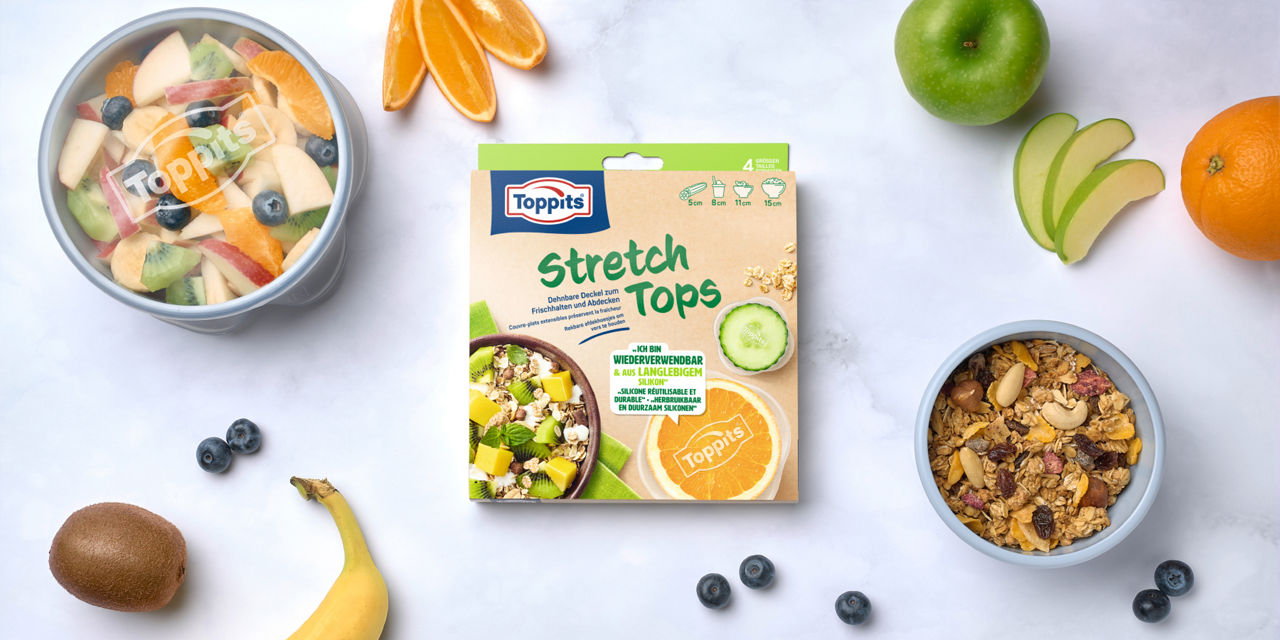 Een verpakking Toppits Stretch Tops ligt tussen verschillende bakjes met vers fruit, muesli, en verspreid fruit zoals banaan, kiwi, appel, sinaasappel en blauwe bessen op een lichte ondergrond.