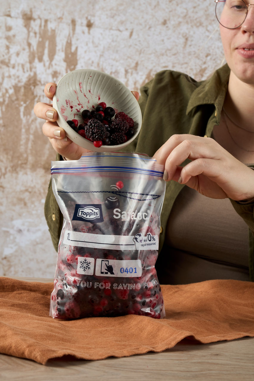 Een vrouw vult een doorzichtige Toppits SafeLoc zak met een mix van bevroren bessen uit een kom, terwijl ze de zak vasthoudt op een tafel met een bruine doek.