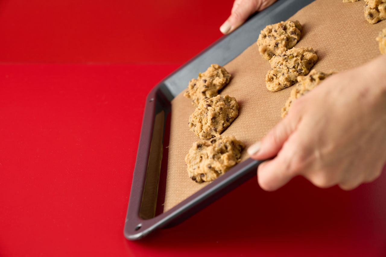 Twee handen houden een bakplaat vast met rauwe koekjesdeeg op een vel bakpapier, klaar om in de oven te worden gebakken, met een rode achtergrond.