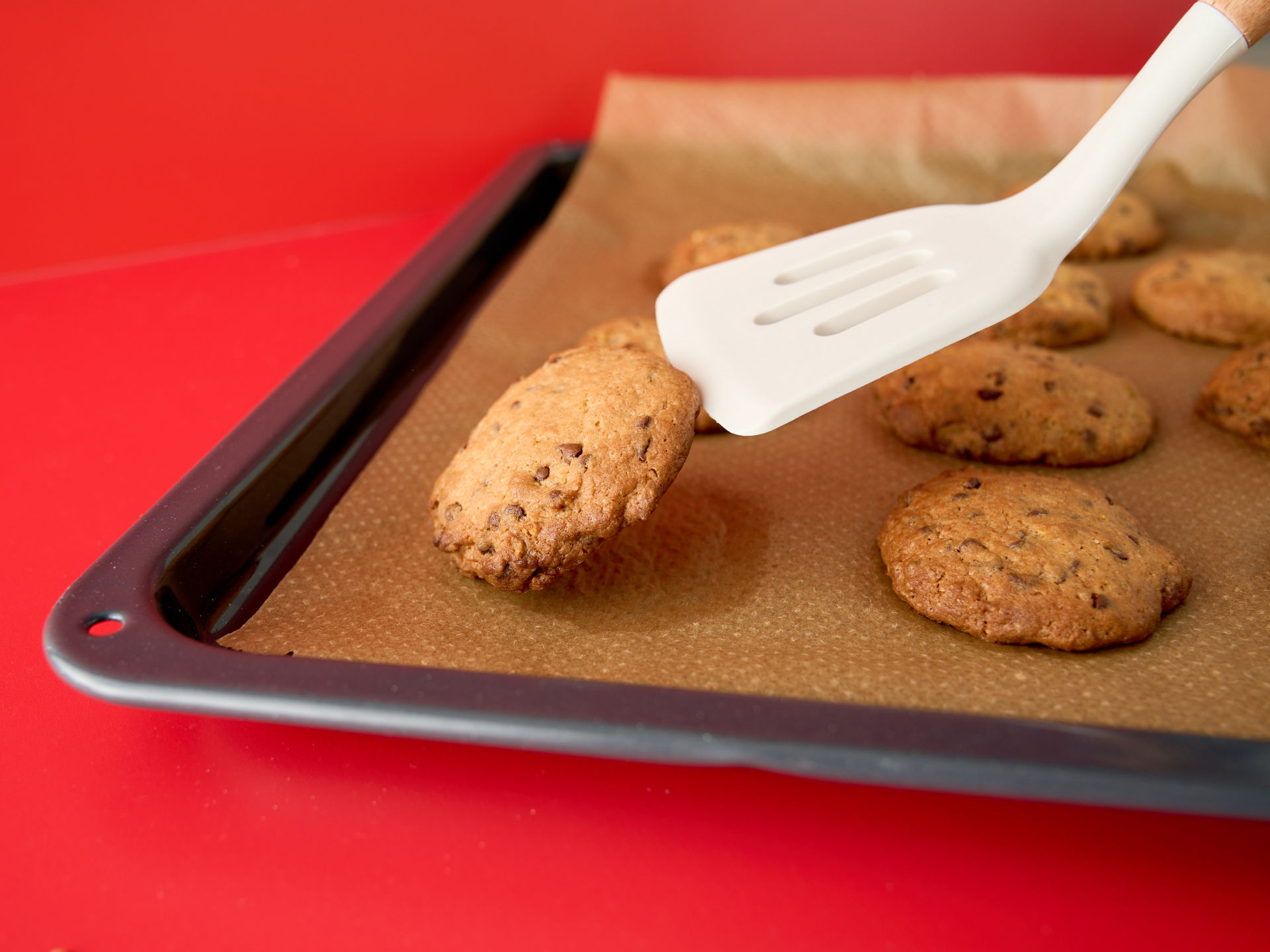 Een witte spatel tilt een versgebakken koekje van een bakplaat met bakpapier, terwijl andere koekjes op de plaat liggen, op een rode ondergrond.
