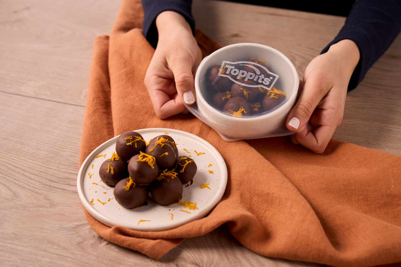 Twee handen plaatsen een Toppits Stretch Top over een schaaltje met chocoladeballetjes met sinaasappelrasp, terwijl er op de voorgrond een bordje met dezelfde lekkernijen ligt.