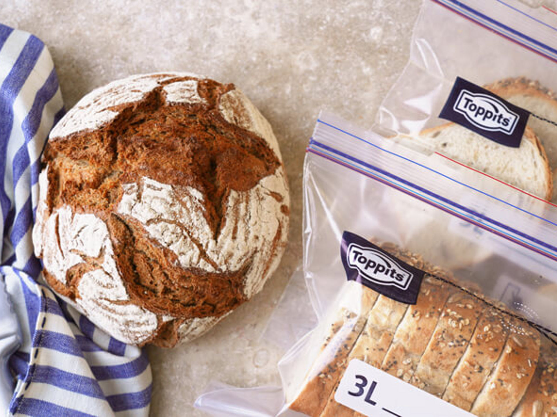 Ein Laib Brot und aufgeschnittenes Brot in zwei Toppits SafeLoc Gefrierbeuteln liegen auf einer hellen Arbeitsfläche neben einem blau-weiß gestreiften Tuch
