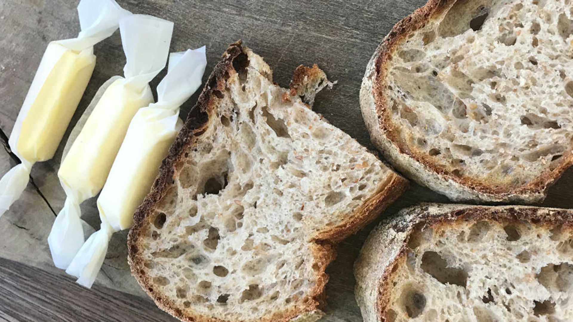 Drei in Toppits Butterbrotpapier gewickelte Butterstücke liegen neben mehreren Scheiben rustikalem Brot auf einem Holztisch