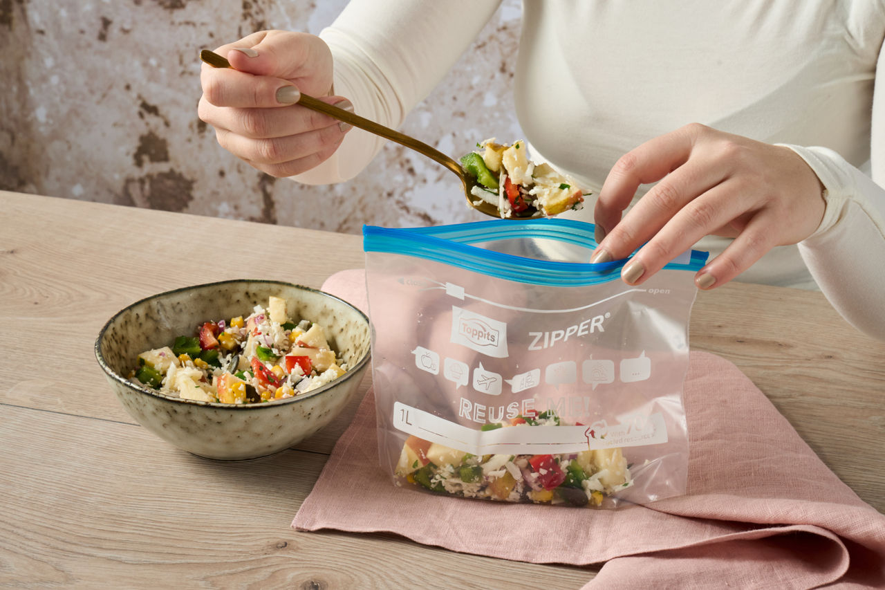 Une personne met de la salade dans un sac Toppits® Zipper® sur une table avec bol et couverts.