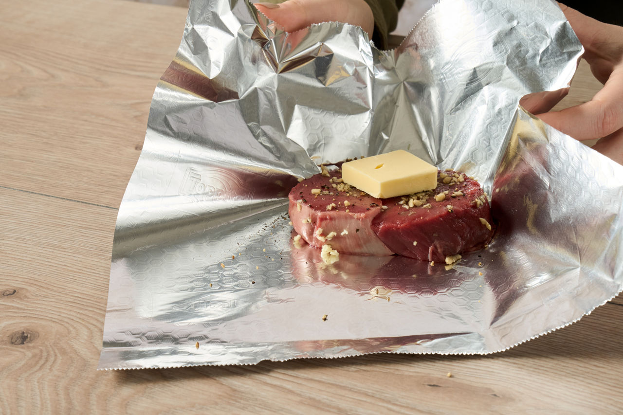 Et stykke kød med smør og krydderier ligger på aluminiumsfolie og pakkes ind