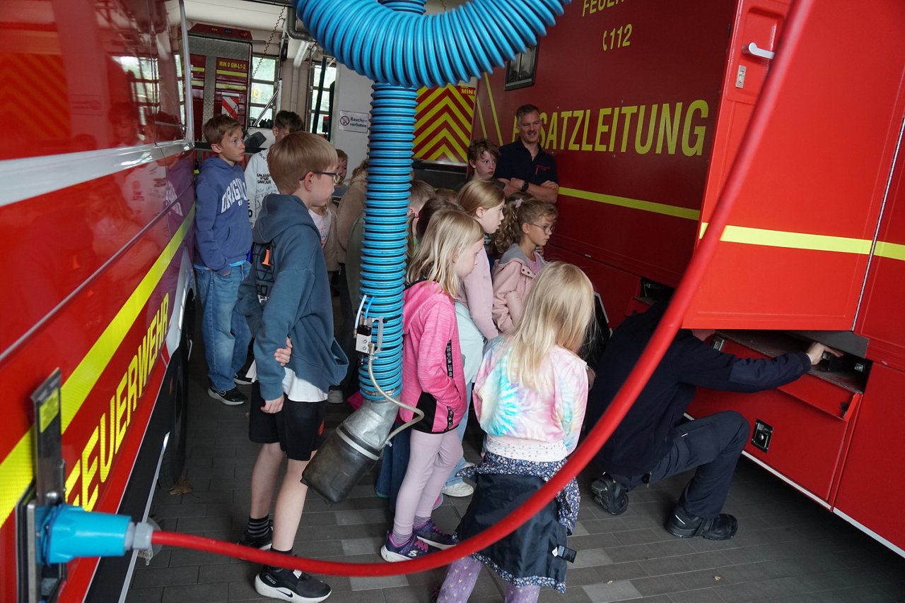 Kinder stehen in einer Fahrzeughalle zwischen zwei roten Feuerwehrfahrzeugen und beobachten einen Feuerwehrmann, der eine Gerätefachklappe öffnet, im Vordergrund ein aufgerolltes blaues Kabel