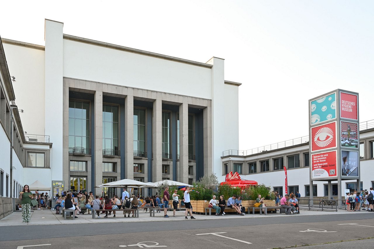 Das Deutsche Hygiene-Museum Dresden am Tag, viele Menschen sitzen und stehen auf dem Vorplatz, im Vordergrund sind Sitzgelegenheiten und ein roter Melitta-Stand zu sehen