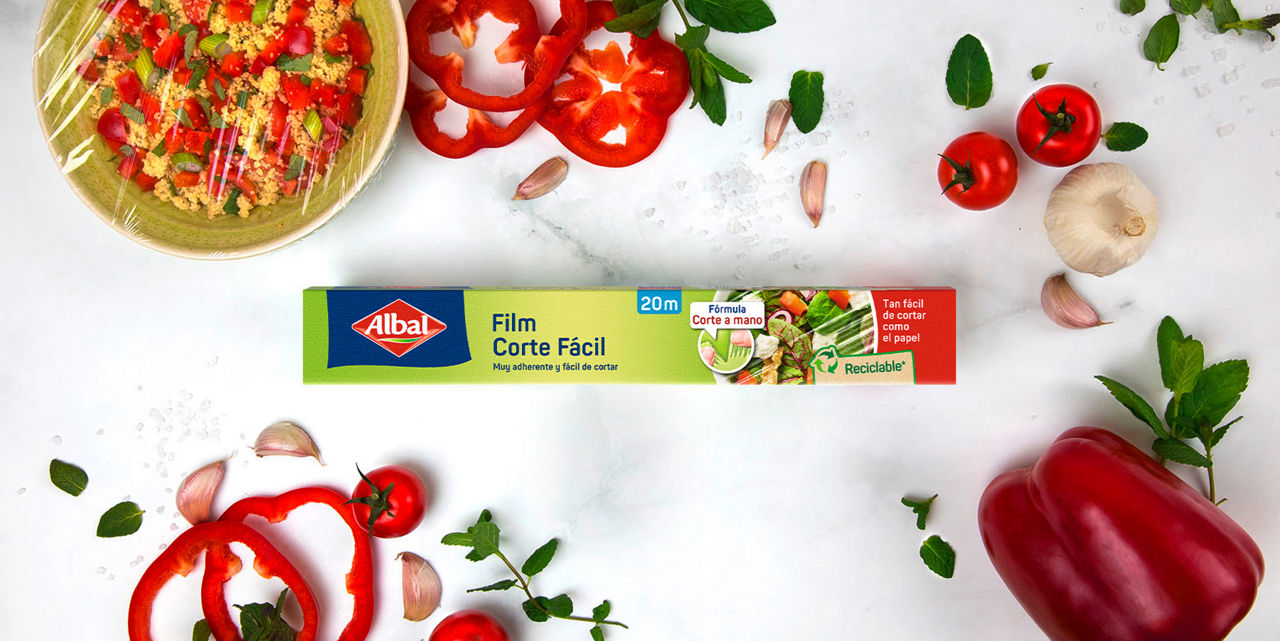 Film corte fácil rodeado de bol de cuscús cubierto con film, pimientos, tomates, ajo y hierbas frescas.