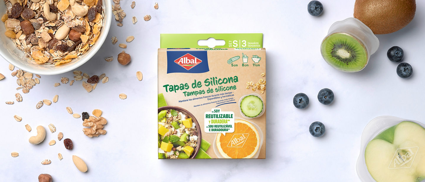 Imagen de una caja de tapas de silicona rodeada de cereales, frutas frescas y recipientes cubiertos con tapas de silicona.