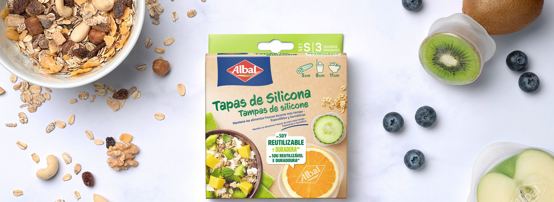 Imagen de una caja de tapas de silicona rodeada de cereales, frutas frescas y recipientes cubiertos con tapas de silicona.