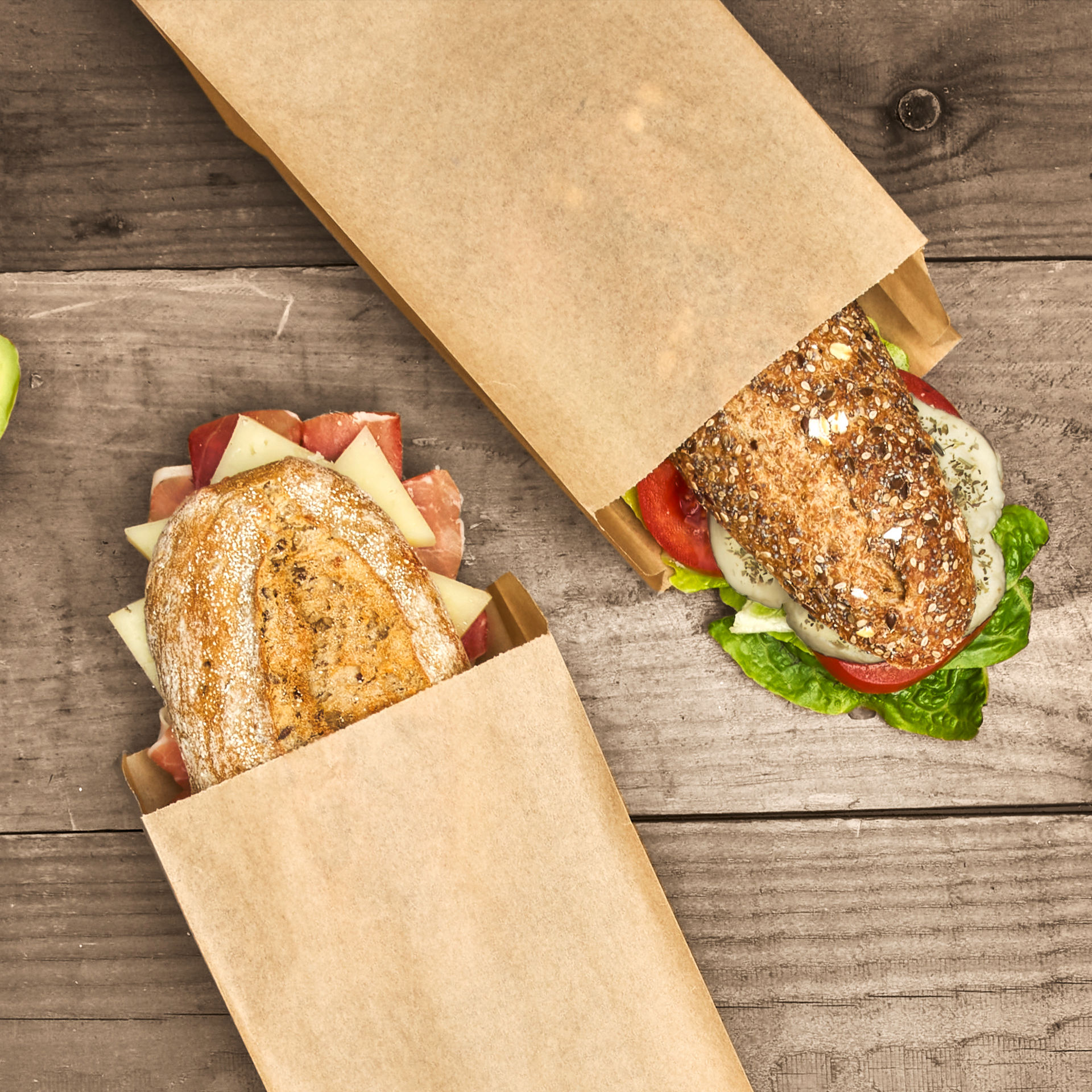 Tres bocadillos variados envueltos en bolsas de papel sobre una mesa de madera oscura.