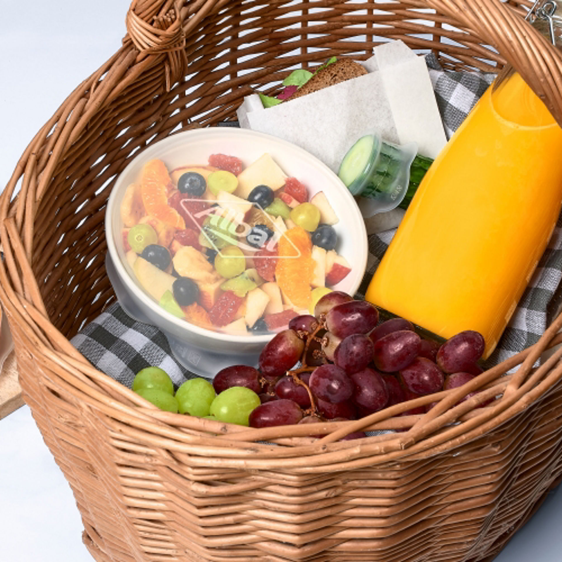 Una cesta de picnic con frutas, zumo, bocadillos y un bol cubierto con tapa de silicona lleno de ensalada de frutas, junto a una tabla con fruta cortada.
