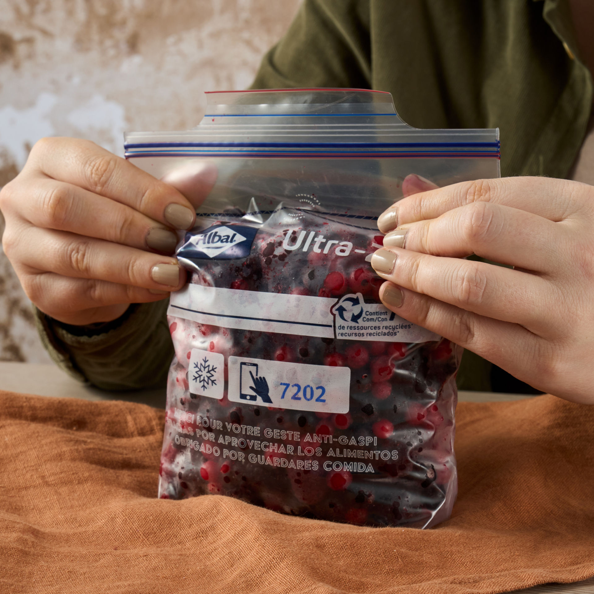 Dos manos sostienen una bolsa de cierre hermético llena de frutos rojos sobre una mesa con paño marrón.