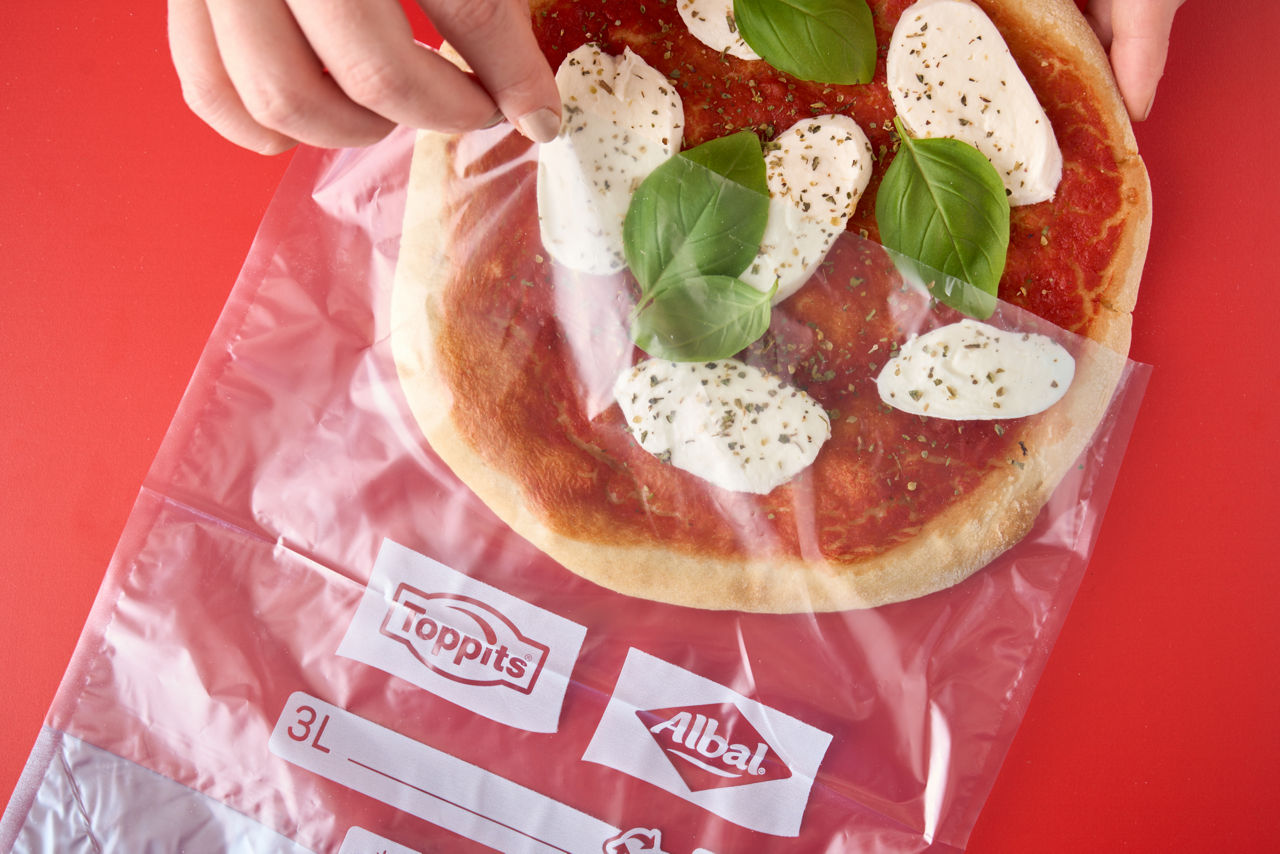 Eine Hand öffnet einen Toppits Gefrierbeutel mit selbstgemachter Pizza auf rotem Hintergrund.