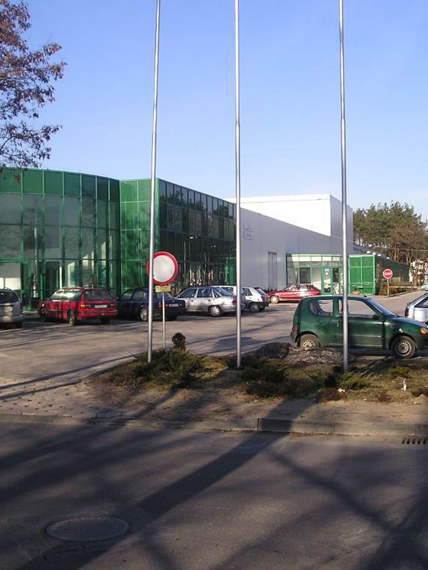 Firmengebäude mit Glasfassade, umgeben von Grünflächen, Parkplätzen und Bäumen, bei Tageslicht. 