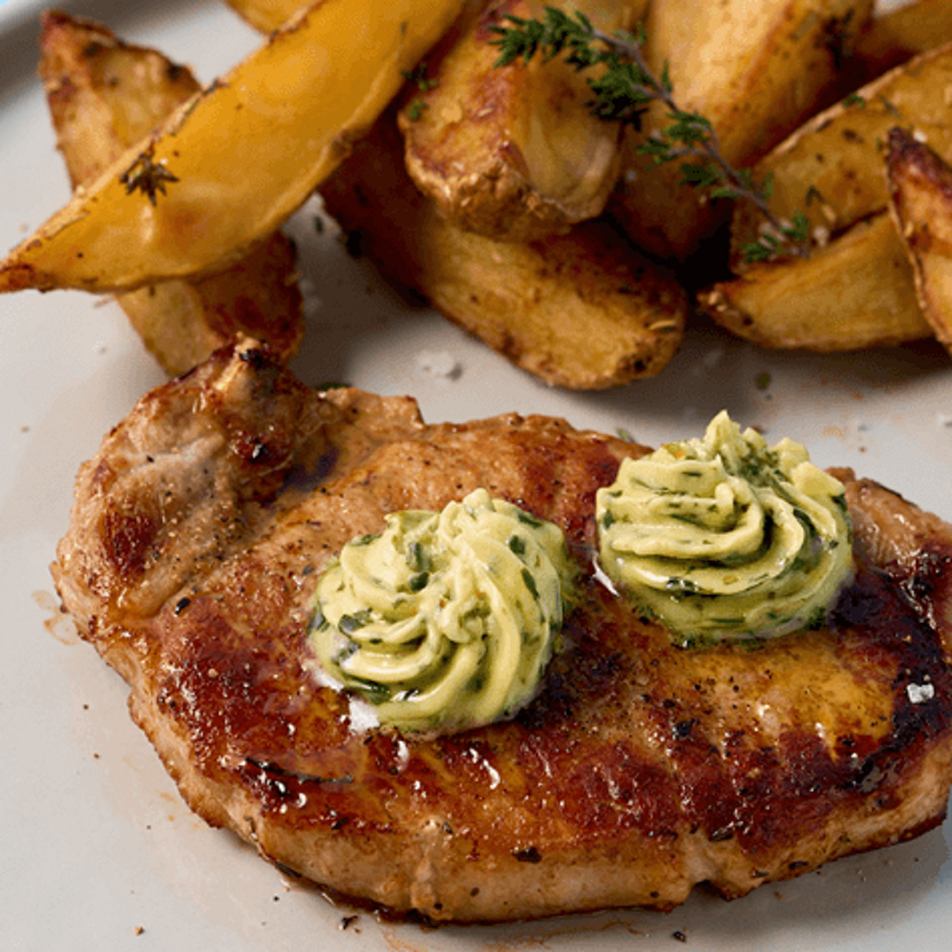 Ein Teller mit einem gebratenen Stück Fleisch, belegt mit zwei Rosetten Kräuterbutter, und knusprigen Kartoffelspalten auf hellblauem Hintergrund