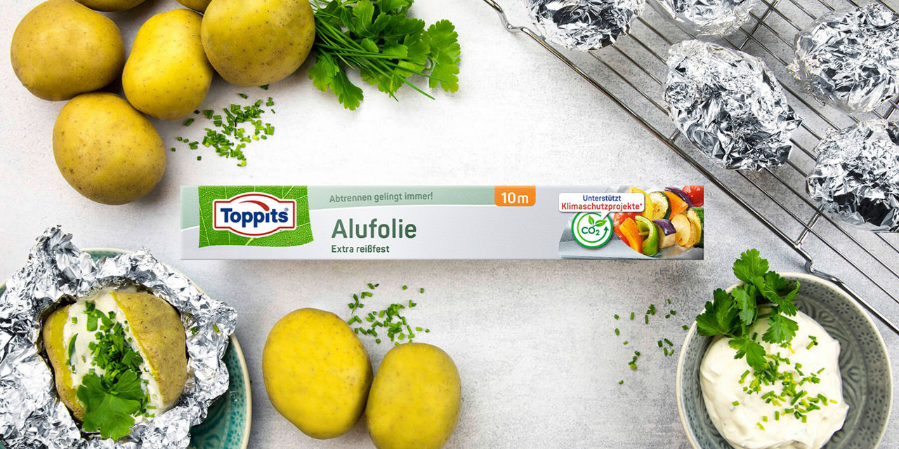 Toppits Alufolie auf einem Tisch mit Kartoffeln, Kräutern, Folienkartoffeln und Kräuterquark; unterstützt Klimaschutzprojekte. 