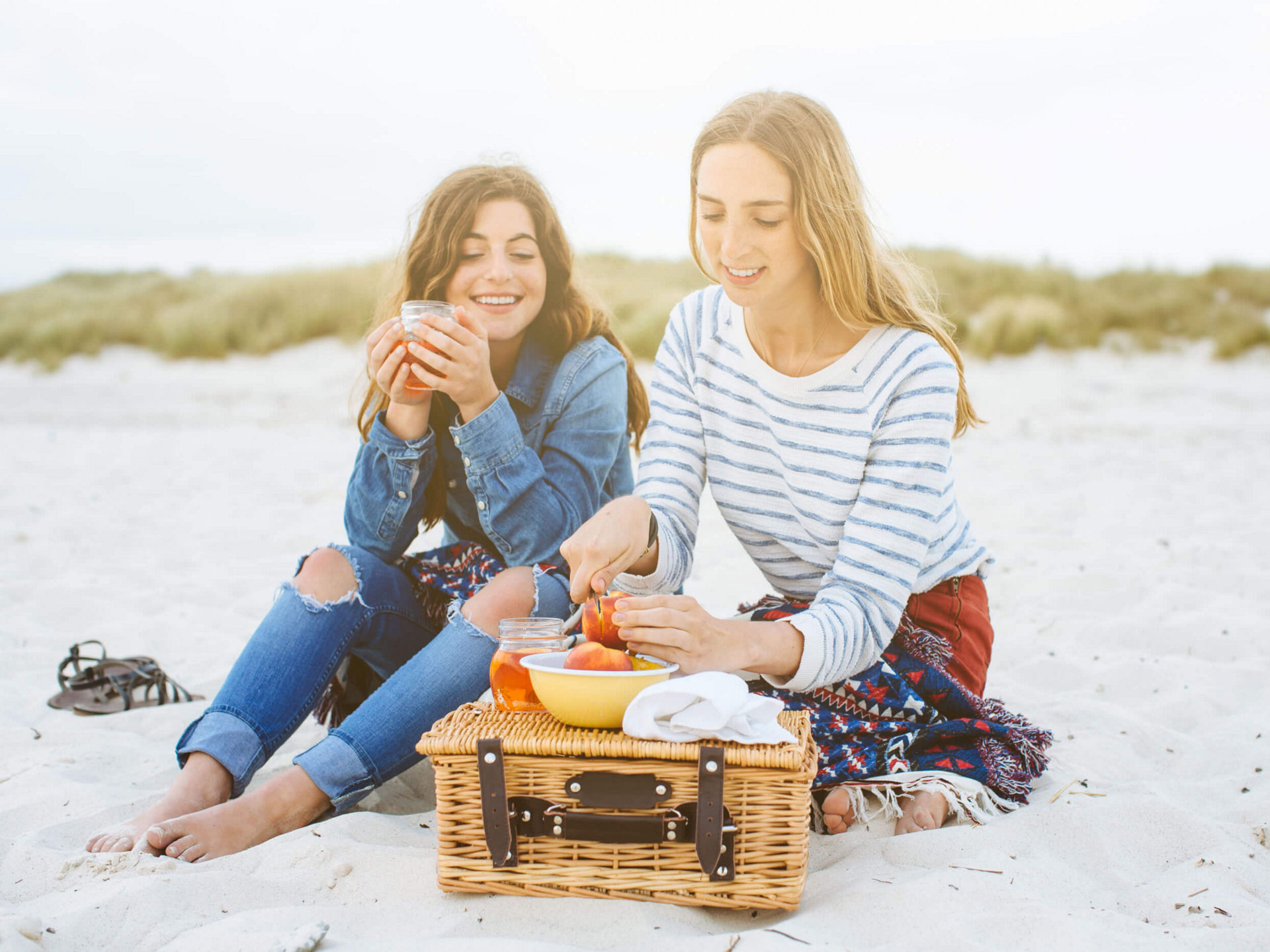 Zwei Personen sitzen am Strand auf einer Decke und genießen ein Picknick mit einem Korb, Obst und Getränken 
