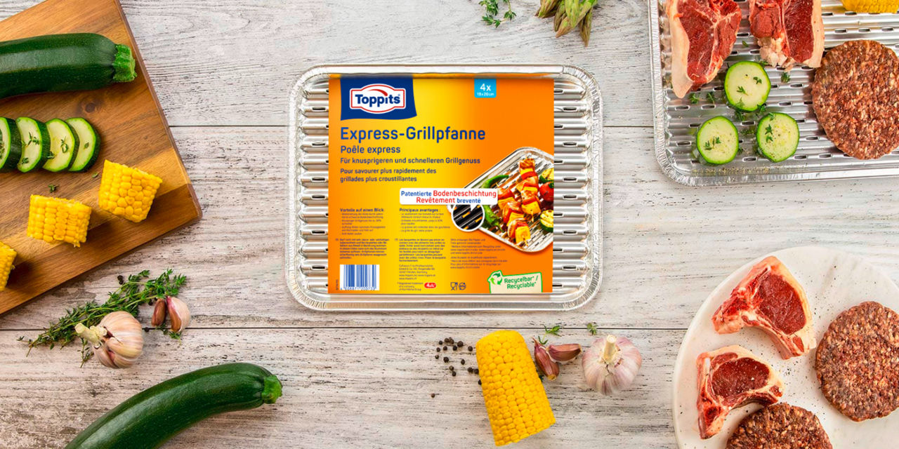 Eine Toppits Express-Grillpfanne steht in der Mitte eines gedeckten Tisches, umgeben von frischem Gemüse wie Zucchini, Mais, Knoblauch und Kräutern sowie rohem Fleisch und Burger-Patties auf Tellern und Grillpfannen