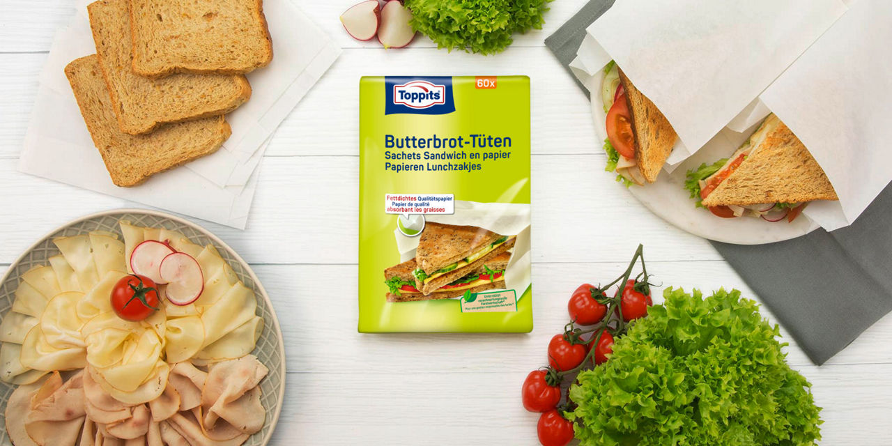 Verpakking van Toppits® sandwichzakjes op een lichte ondergrond, met daarnaast belegde boterhammen, verse groenten, sneetjes brood en een bord met vleeswaren 