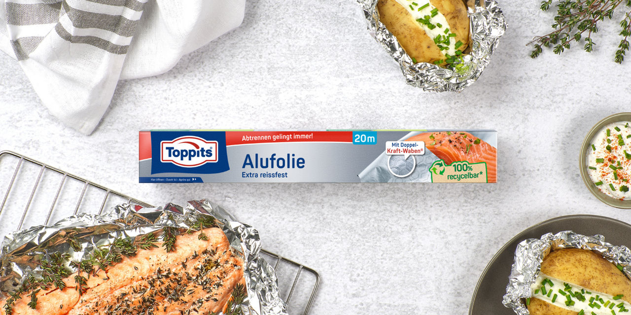  Eine Packung Toppits Alufolie (20 Meter, extra reißfest, 100% recycelbar) liegt auf einem hellen Tisch, umgeben von in Alufolie gebackenem Lachs mit Kräutern und Folienkartoffeln mit Kräuterquark.
