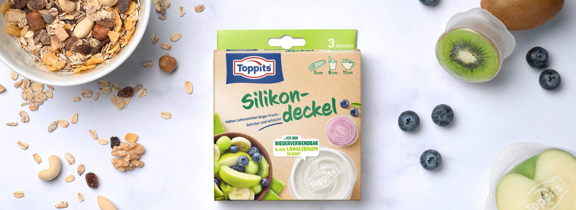 Packung Toppits Silikondeckel auf einem Tisch, daneben Müsli, Nüsse, eine Kiwi, Blaubeeren und ein Apfelstück mit Silikondeckel