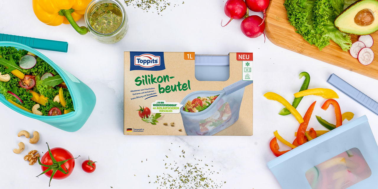 Toppits Silikonbeutel in Verpackung auf einem Tisch, daneben frischer Salat, geschnittenes Gemüse, Nüsse und ein Glas Dressing