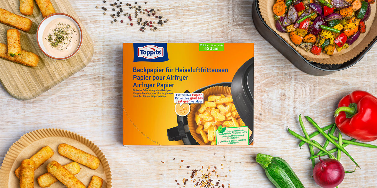 Eine Packung Toppits Backpapier für Heißluftfritteusen (Airfryer Papier, 20 Stück, Ø 20 cm) liegt auf einem Holztisch, umgeben von panierten Sticks, einer Schale mit Dip, Gemüse und einer Heißluftfritteuse mit buntem Ofengemüse.