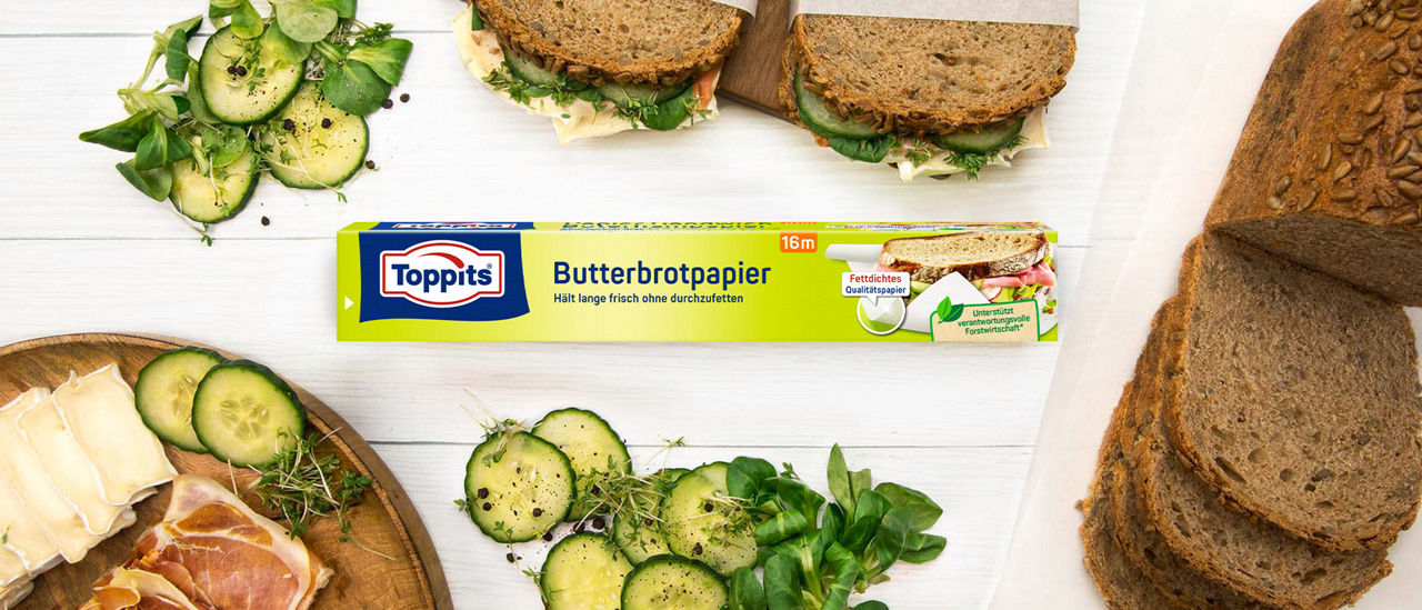Packung Toppits Butterbrotpapier, umgeben von belegten Broten, Gurkenscheiben, Salat, Käse und Brotlaib