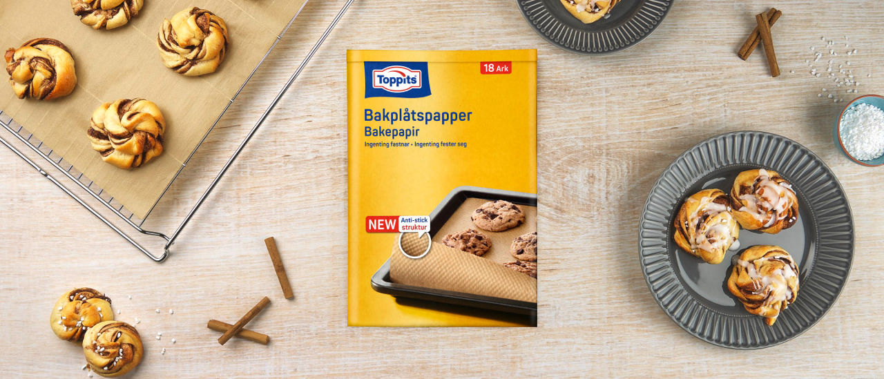 Headerbild med Toppits® bakplåtspapper-produktförpackning, bakplåtspapper och nybakat fikabröd på ljus bakgrund