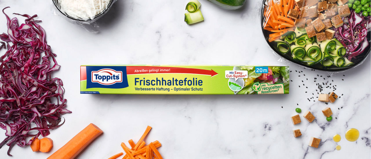 Packung Toppits Frischhaltefolie 20m liegt auf einer weißen Marmorfläche, umgeben von geschnittenem Rotkohl, Karotten, Zucchini, Erbsen, Tofu und einer Bowl mit frischem Gemüse, teilweise mit Frischhaltefolie abgedeckt.