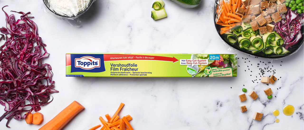 Een verpakking Toppits vershoudfolie ligt centraal op een marmeren ondergrond, omringd door gesneden rode kool, wortels, komkommer en een saladebowl die is afgedekt met folie.