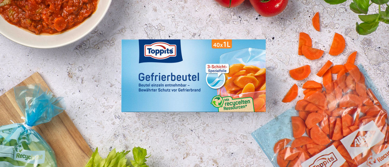 Eine Packung Toppits Gefrierbeutel liegt auf einem Tisch, umgeben von geschnittenen Karotten, frischen Tomaten, Kräutern und einem Beutel mit Gemüse 