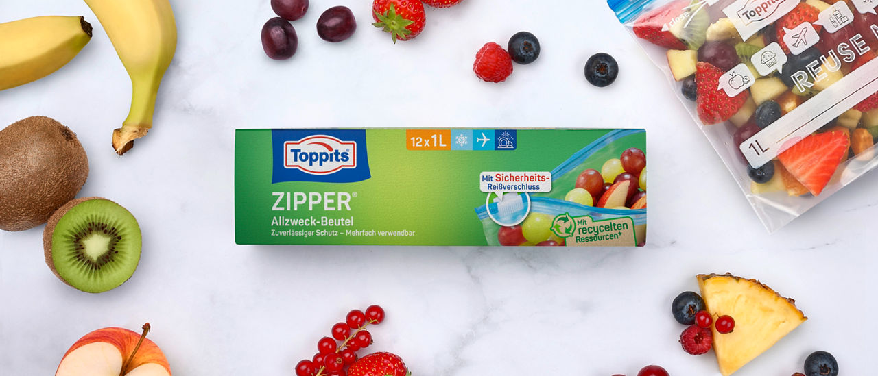 Packung Toppits Zipper Allzweck-Beutel und ein gefüllter Zipper-Beutel, umgeben von frischem Obst. 
