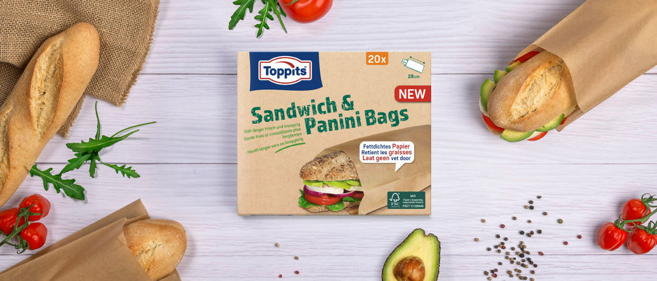 Eine Packung Toppits Sandwich & Panini Bags liegt auf einem Tisch, umgeben von belegten Brötchen, Tomaten, Avocado und Rucola. 