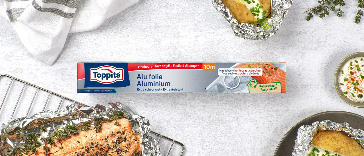 Een pak Toppits-aluminiumfolie ligt in het midden van een tafel, omringd door gerechten zoals in folie verpakte aardappelen en zalm, samen met verse kruiden en een kommetje saus.