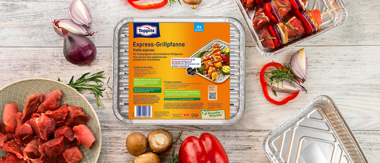 Eine Toppits Express-Grillpfanne steht in der Mitte eines gedeckten Tisches, umgeben von frischem Gemüse wie Zucchini, Mais, Knoblauch und Kräutern sowie rohem Fleisch und Burger-Patties auf Tellern und Grillpfannen