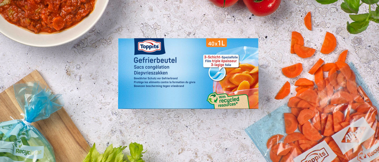 Eine Packung Toppits Gefrierbeutel liegt auf einem Tisch, umgeben von geschnittenen Karotten, frischen Tomaten, Kräutern und einem Beutel mit Gemüse