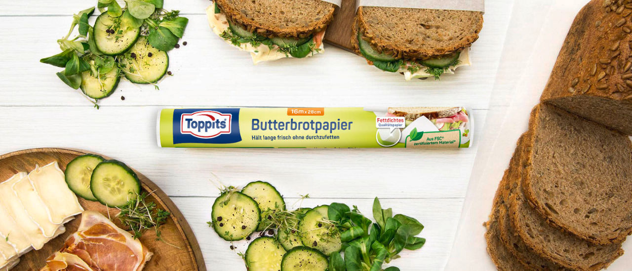  Toppits Butterbrotpapier mit belegten Broten, frischen Zutaten und Vollkornbrot auf Holzbrett.