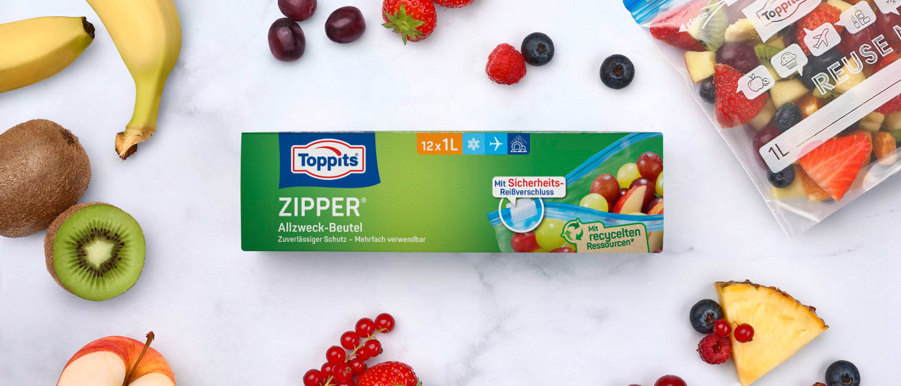 Packung Toppits Zipper Allzweck-Beutel und ein gefüllter Zipper-Beutel, umgeben von frischem Obst. 