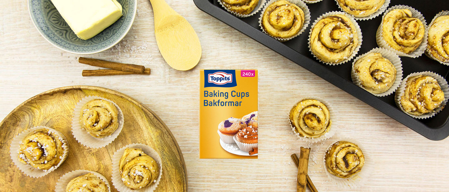 Verpakking Toppits® Baking Cups op een licht werkblad, omringd door gebakken kaneelbroodjes of muffins in bakvormen, op borden en met bakgere  