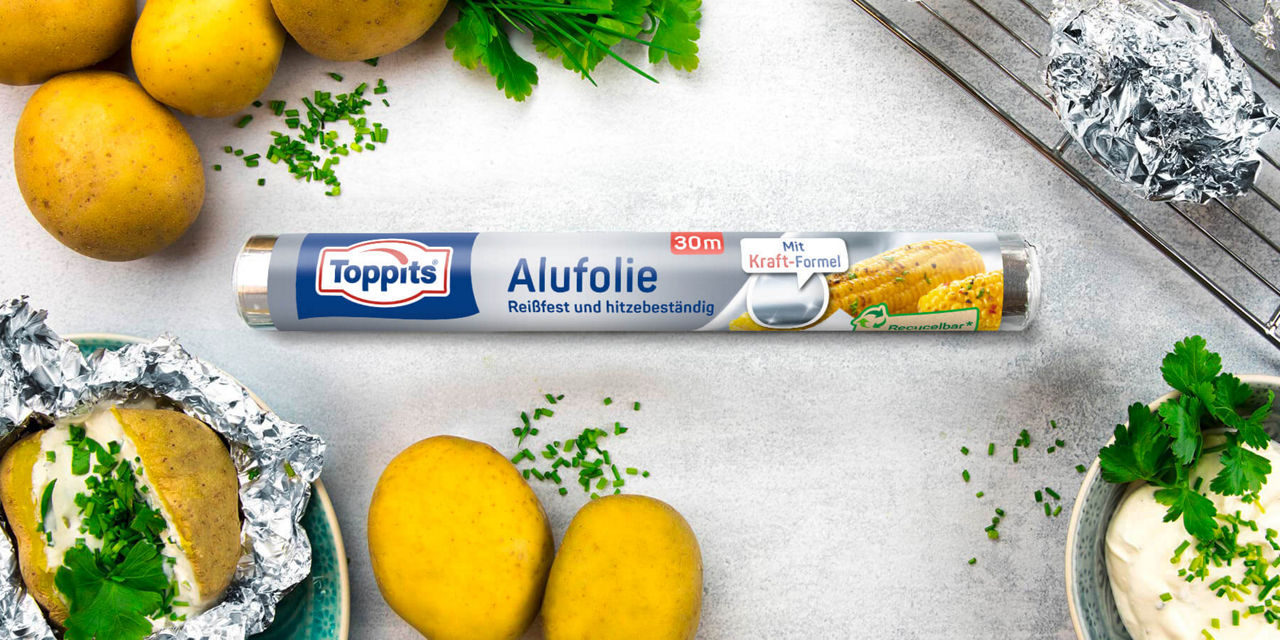 Toppits Alufolie-Rolle liegt zwischen Kartoffeln, Kräutern und Alufolie auf einem Tisch. 