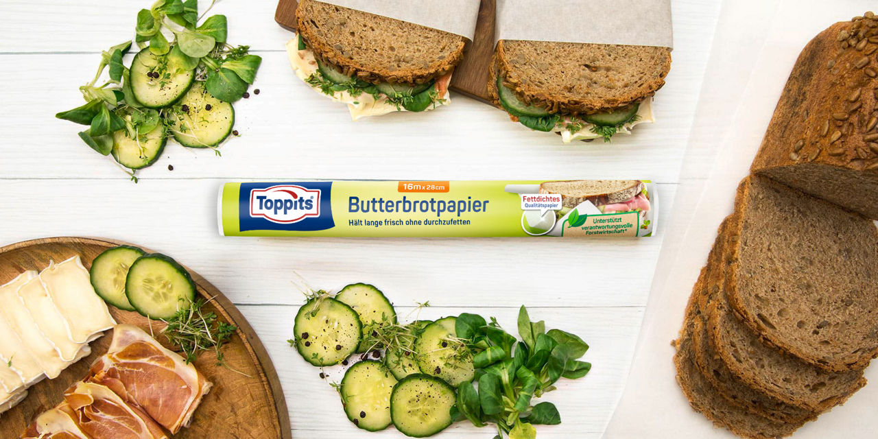 Toppits Butterbrotpapier mit belegten Broten, frischen Zutaten und Vollkornbrot auf Holzbrett.