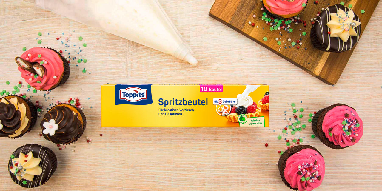 Eine Packung Toppits Spritzbeutel liegt auf einem Holztisch, umgeben von dekorierten Cupcakes mit rosa und schokoladenfarbener Glasur. Ein gefüllter Spritzbeutel befindet sich darüber, während bunte Zuckerstreusel verstreut sind.