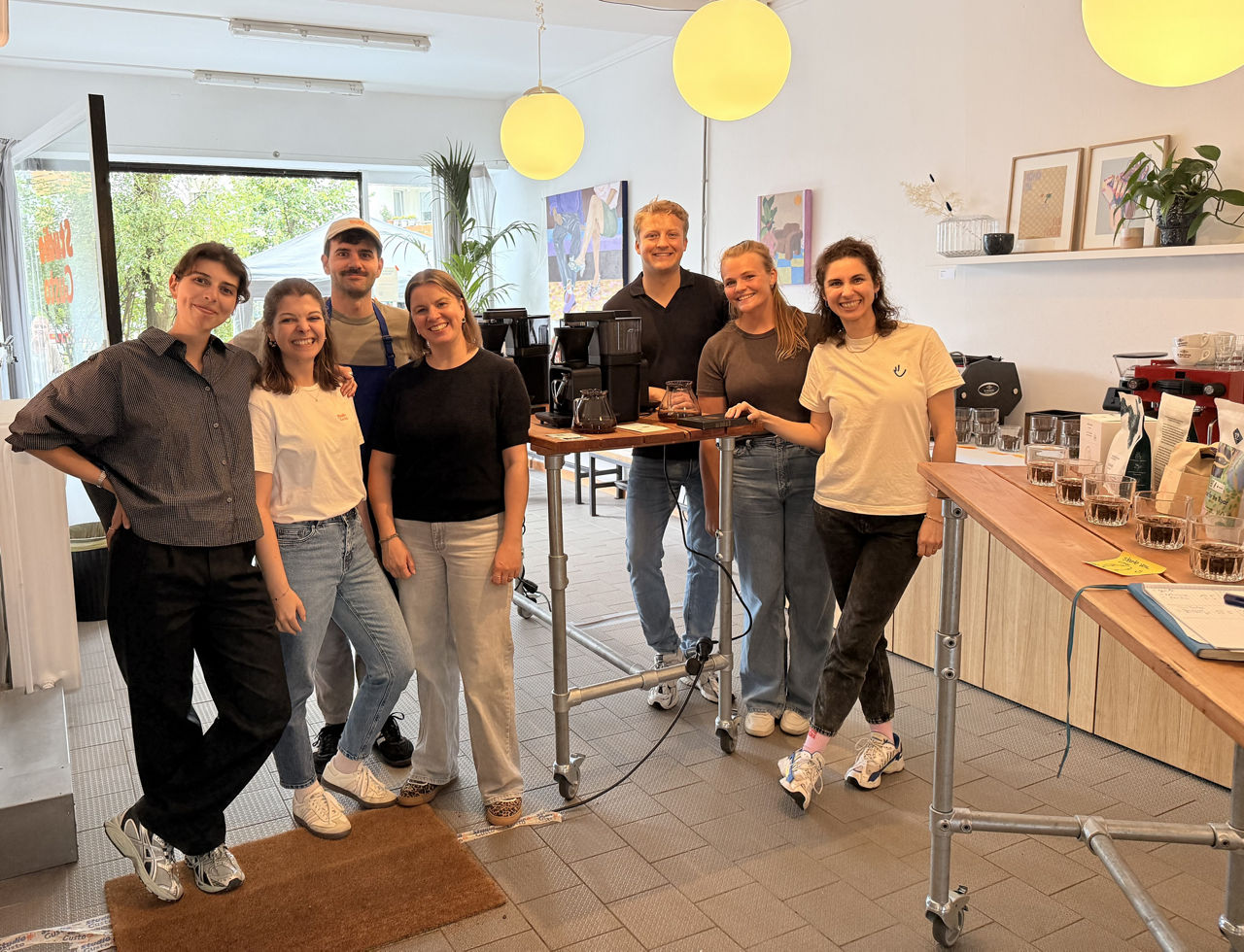 Gruppe von sieben Personen steht lächelnd nebeneinander in einem hellen Raum mit Kaffeemaschinen, Regalen und gelben Hängelampen im Hintergrund. 
