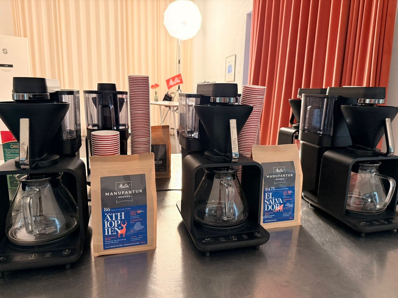 Mehrere schwarze Kaffeemaschinen stehen nebeneinander auf einem Tisch vor einem roten Vorhang; daneben Bücher und Zubehör.