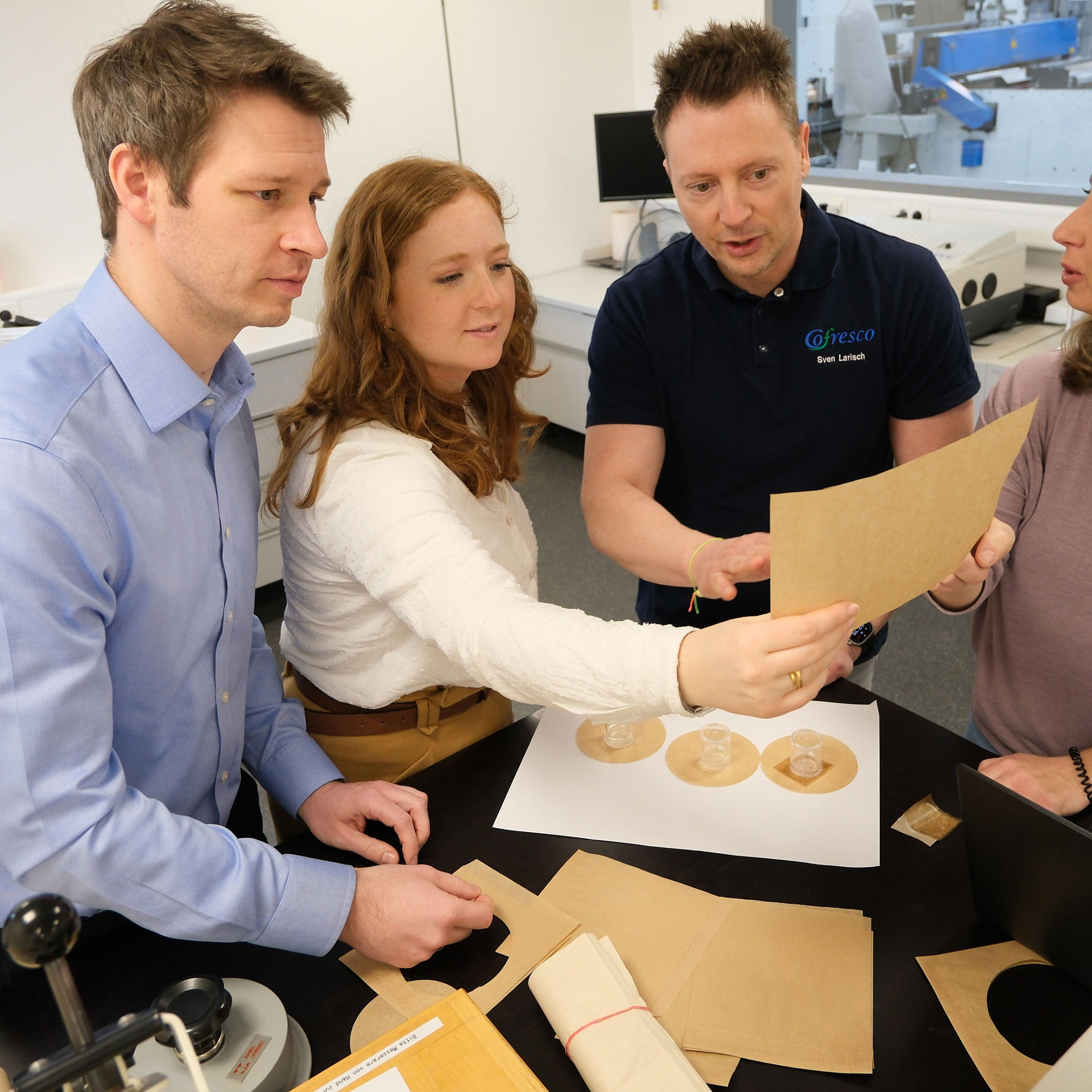 Vier Personen stehen in einem Labor und begutachten gemeinsam verschiedene Papierproben und runde Zuschnitte auf einem Tisch. Auf dem Tisch liegen zudem Laborgeräte und Unterlagen. Im Hintergrund sind technische Geräte und Monitore zu sehen