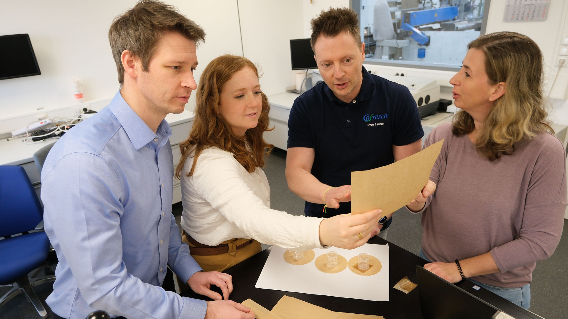 Vier Personen stehen in einem Labor und begutachten gemeinsam verschiedene Papierproben und runde Zuschnitte auf einem Tisch. Auf dem Tisch liegen zudem Laborgeräte und Unterlagen. Im Hintergrund sind technische Geräte und Monitore zu sehen