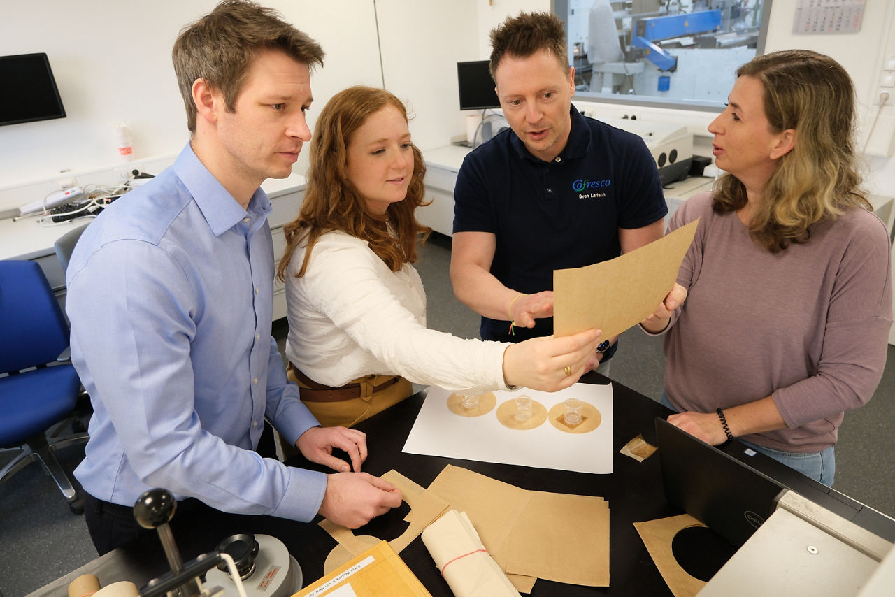 Vier Personen stehen in einem Labor und begutachten gemeinsam verschiedene Papierproben und runde Zuschnitte auf einem Tisch. Auf dem Tisch liegen zudem Laborgeräte und Unterlagen. Im Hintergrund sind technische Geräte und Monitore zu sehen