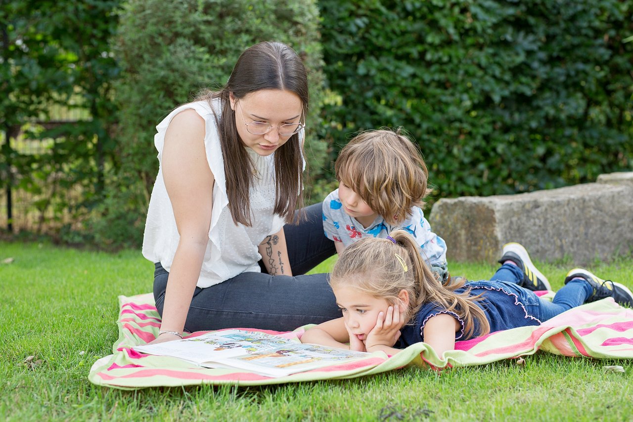 Woman with dark hair sitting on a blanket outside on the lawn together with two children. They read a book together.